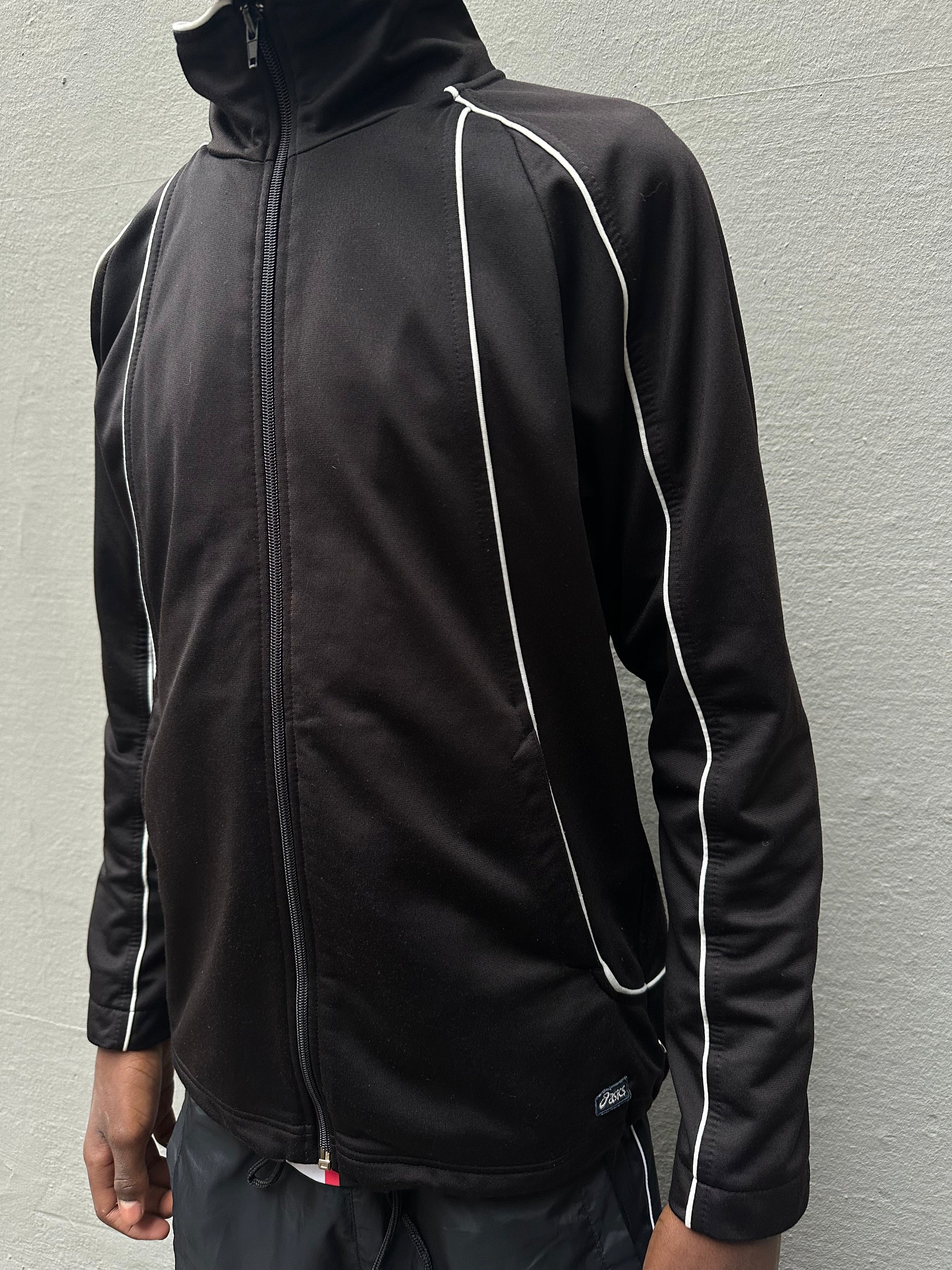 Tragebild schwarze ASICS Track Jacket  vor grauem Boden von vorne.