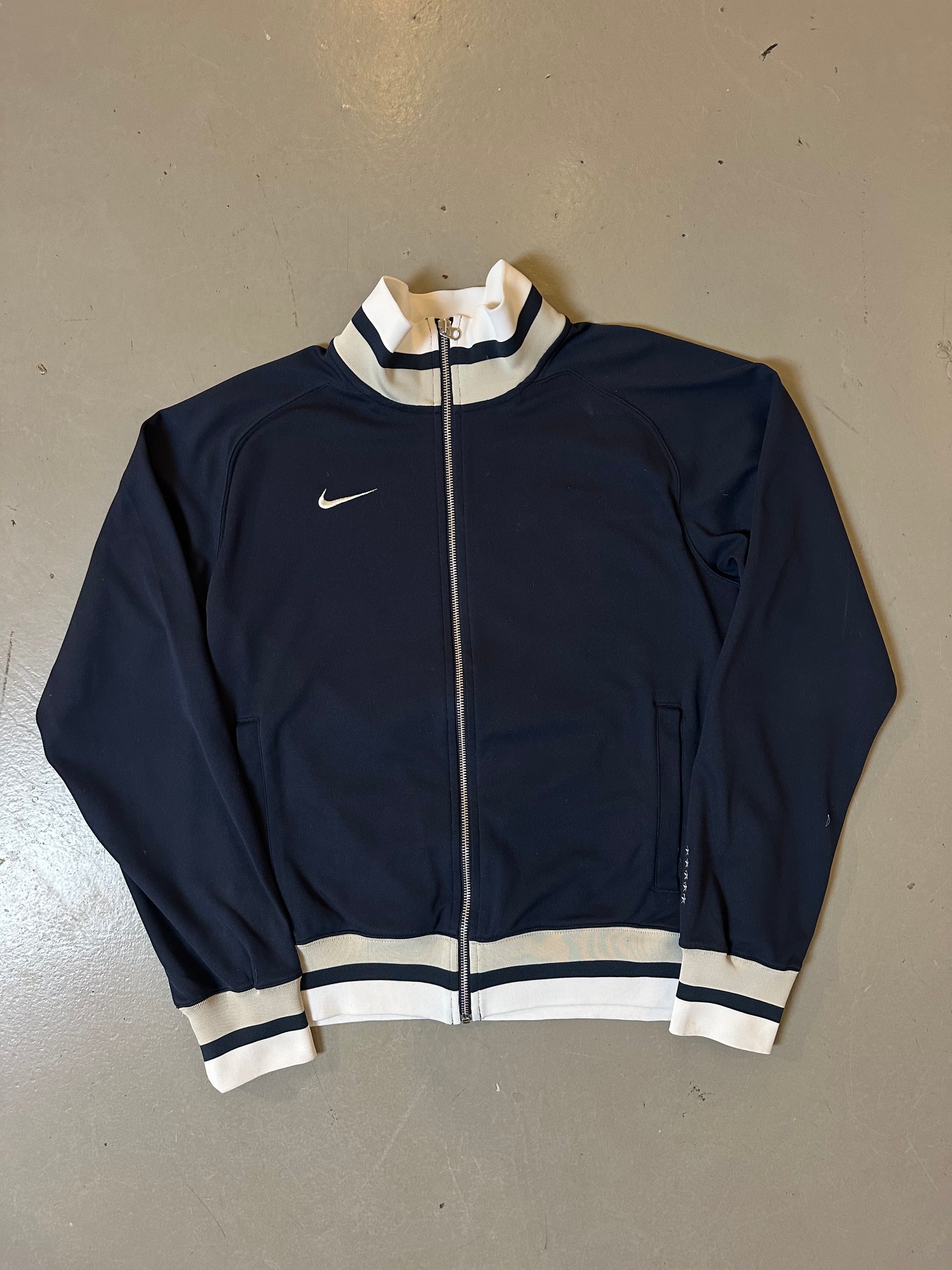 Zu sehen ist eine blaue Trackjacke von Nike it weißem Bund an kragen und Ärmel 