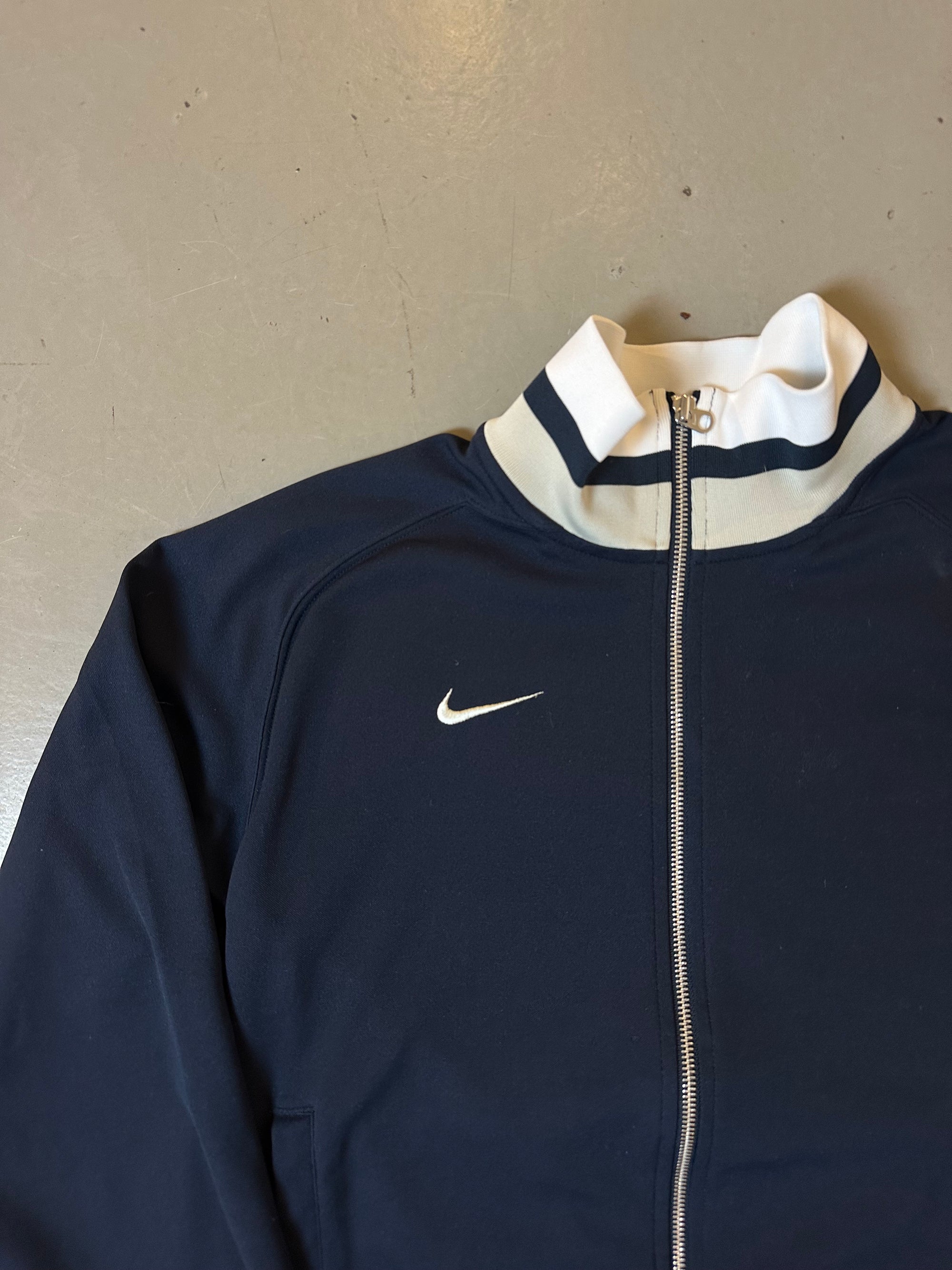 Zu sehen ist eine blaue Trackjacke von Nike it weißem Bund an kragen und Ärmel 