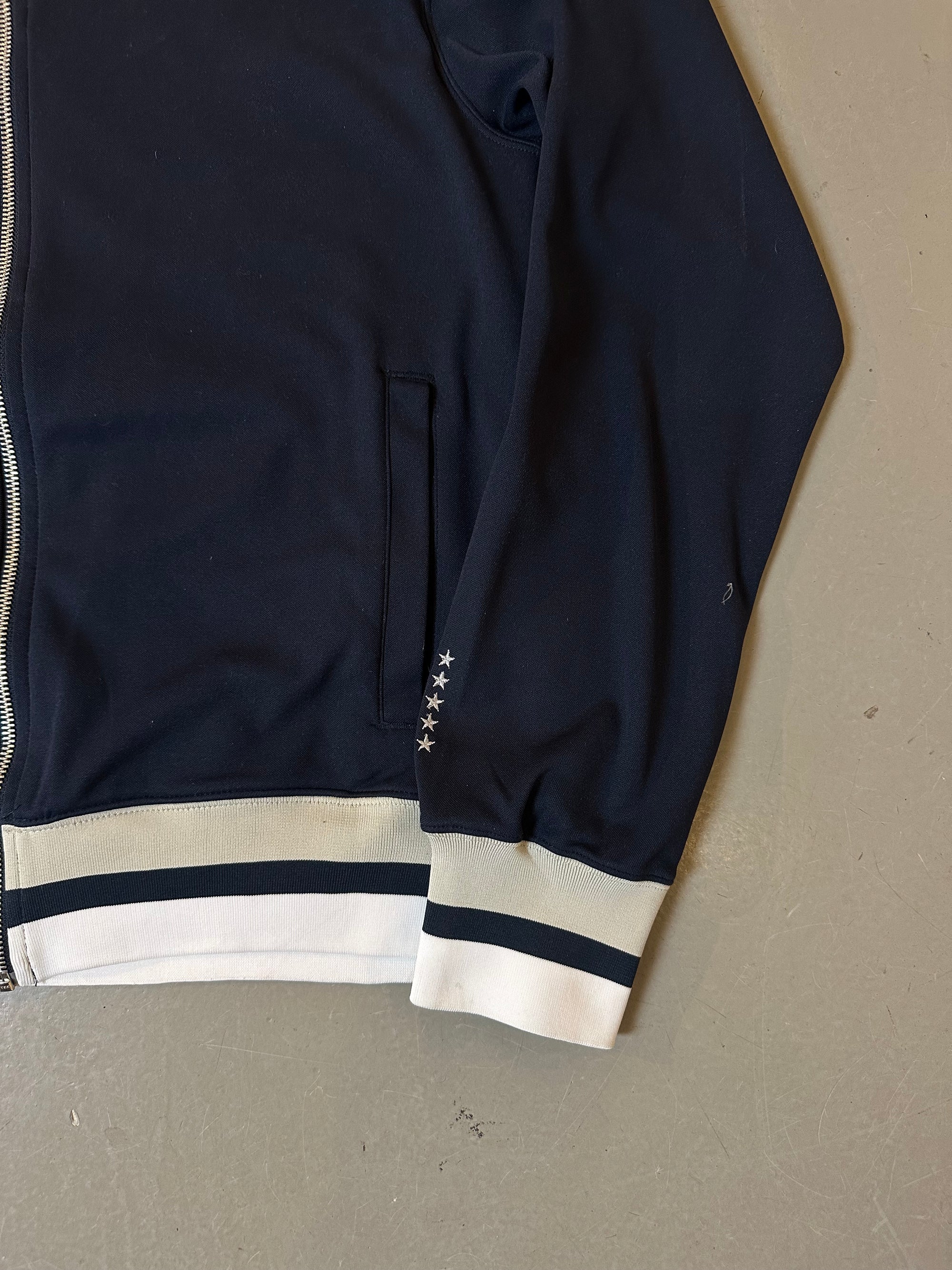 Zu sehen ist eine blaue Trackjacke von Nike it weißem Bund an kragen und Ärmel 