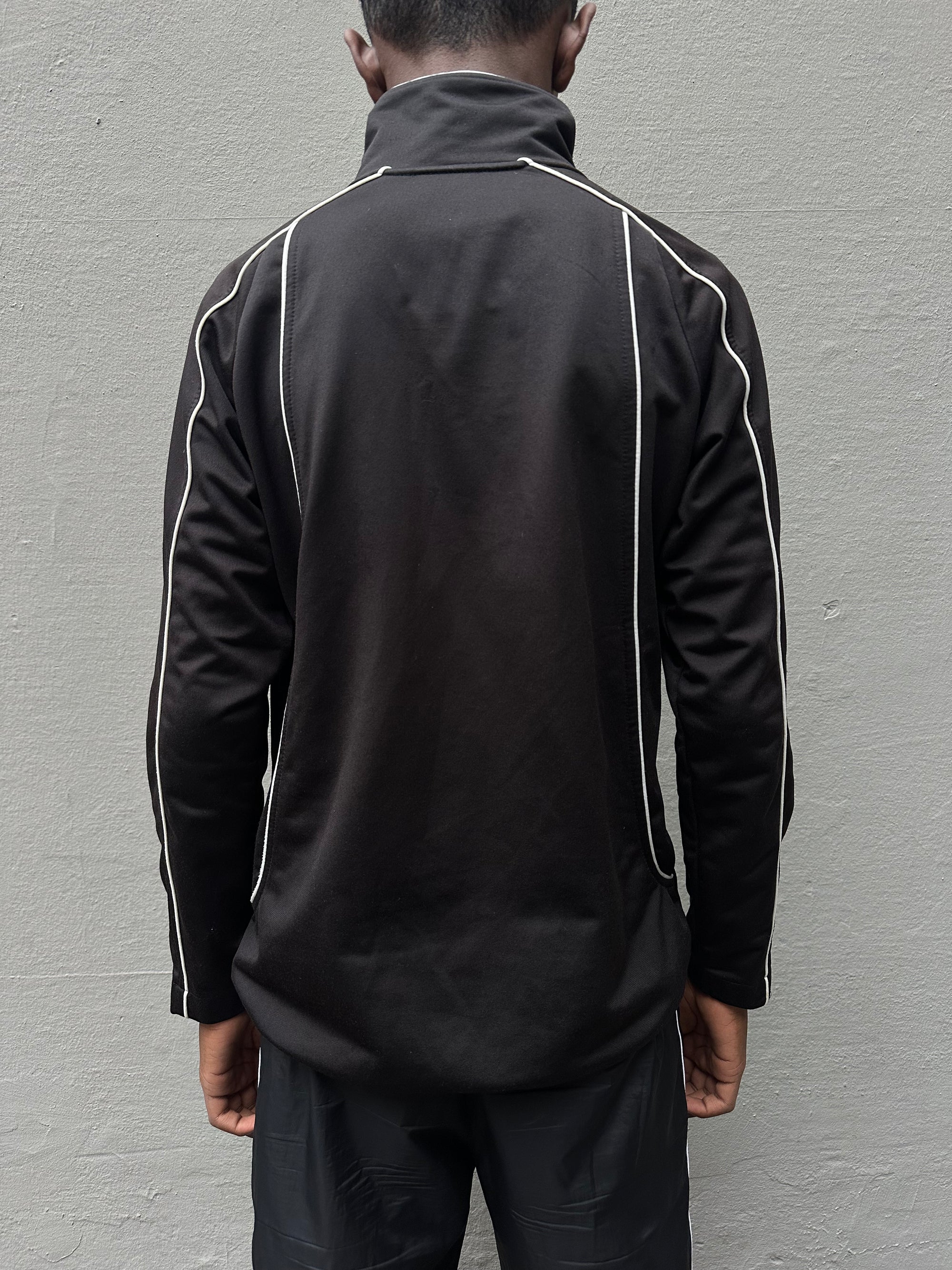 Tragebild schwarze ASICS Track Jacket vor grauer Wand von hinten.