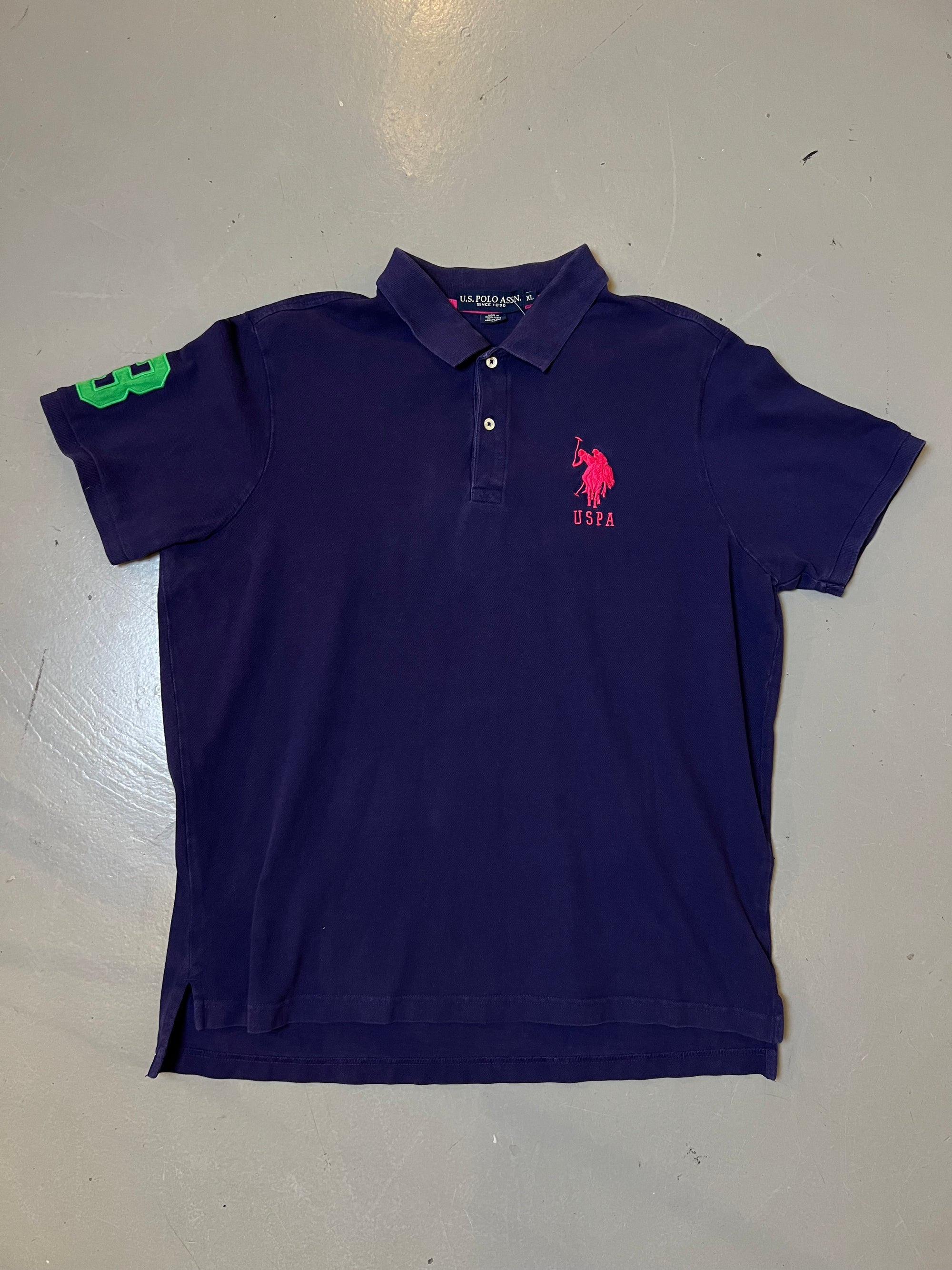 Zu sehen ist Ring lila Polo Shirt von U.S. Polo Assn. mit dem pinken Logo auf der Brust und einer grünen 3 auf dem Ärmel 